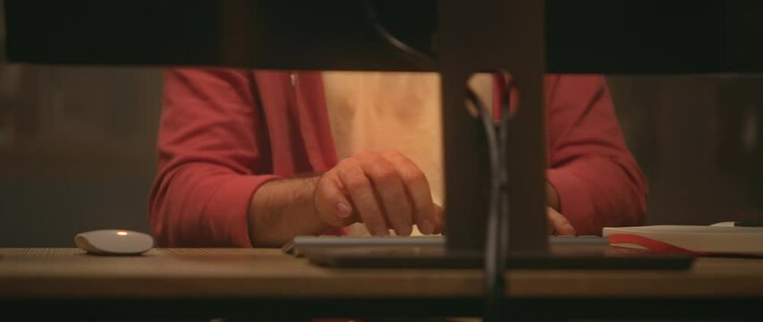 Tracking moving around shot of unrecognizable Caucasian young man typing on keyboard of computer and writing down notes in notebook. Modern office in evening. Cinematic. Male hands. Parallax effect