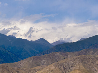 Fototapeta premium A picturesque view of majestic mountains with varying shades of green and brown, under a sky filled with fluffy clouds. The rugged terrain contrasts beautifully with the serene sky above.