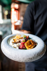 A close-up of a gourmet dish held by a person, featuring vibrant colors and textures. The dish includes various fresh ingredients, artistically arranged, making it visually appealing and appetizing