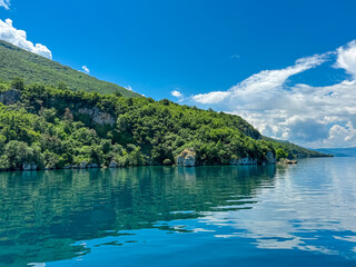 Obraz premium Serene lakeside scene along the shoreline of Lake Ohrid, North Macedonia. Mountain range of National Park Galicica in the distance. Slopes and hills covered with lush green vegetation and forest