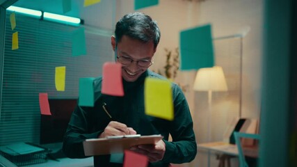 Asian project manager is smiling while brainstorming, taking notes, and organizing tasks using sticky notes on a glass wall in the office at night