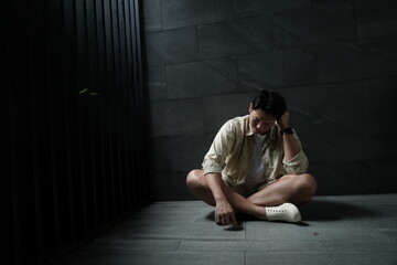 Asian man in shorts sits on the floor in a dark corner outside his house feeling bored, anxious, and lonely.