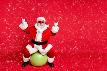 Full length body size view of his he nice cheerful cheery glad fat bearded Santa working out sitting on pilates ball showing horn sign heavy metal isolated over bright vivid shine red background