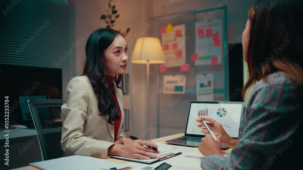 Poster two businesswomen work late in the office, analyzing data on a laptop and digital tablet while discu