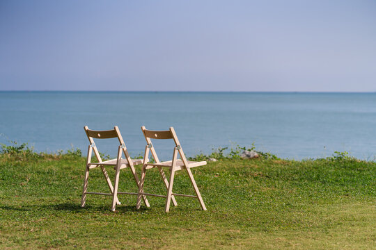 Wooden chairs placed on a grassland, facing the sea and sky in background.