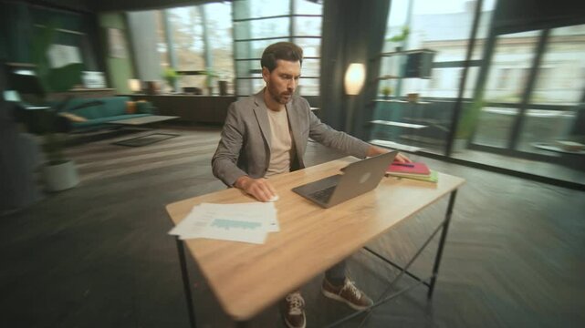 Shot of serious young Caucasian man sitting at desk and holding pink notebook in hands. 360 degree time- lapse. Footage of good-looking businessman typing on keyboard of laptop in spacious office