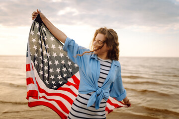 Young woman holding national American flag walking ocean beach. America Independence Day concept....