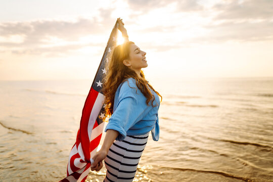 Young woman holding national American flag walking ocean beach. America Independence Day concept. 4th of July.