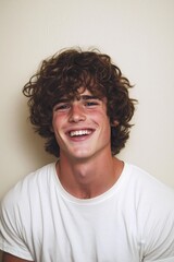 Obraz premium Cheerful Young Man with Curly Hair in White T-Shirt, Warm Smile Portrait on Textured Off-White Background - Genuine Happiness Concept
