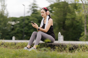 Fototapeta premium Woman Taking a Break: A young, athletic woman in sportswear takes a break from her workout, listening to music through her headphones while using her smartphone.