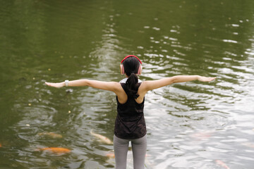 Fototapeta premium Finding Peace by the Pond: A woman stands by a pond, arms outstretched, seeking solace in the tranquil natural surroundings. The water is teeming with fish, a symbol of abundance and life.