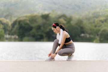 Ready to Run: A young woman in athletic wear prepares for her workout, tying her shoes by a serene lake with lush green foliage. She's wearing headphones and a towel.