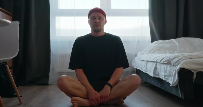 A single man enjoys his free time, meditates sitting on the floor in his bedroom and does breathing exercises. A millennial man in a black T-shirt meditates next to the bed. High quality 4k footage