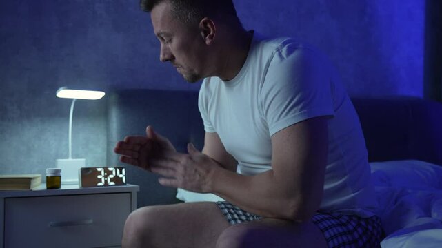 Young man suffering from insomnia while sitting in bed  at night. Depressed man having headache or migraine, holding head with hands