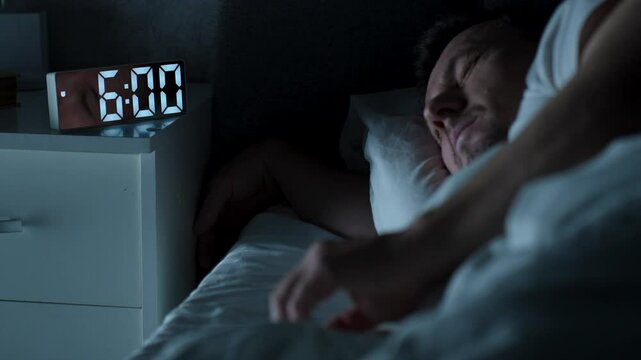Young man sleeping in bed pressing snooze button on alarm clock at six o'clock morning in bedroom, selective focus