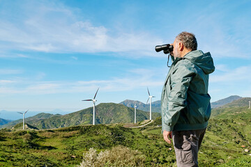 Male tourist using binoculars in front of wind turbine in wind farm with mountain landscape view.