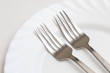 Silver cutlery background. White tablecloth with ceramic plate. Two forks elegant dishware. Dining at restaurant. Table arrangement background. Clean shiny metal silverware.