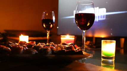 Romantic dinner. In the foreground is a plate with sushi or rolls. Next to it are two glasses of red wine. In the background are three candles.The atmosphere is cozy and warm.