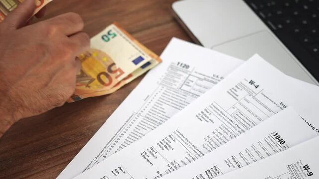 Businessman counts out money by transferring euro banknotes from hands to wooden table, person places bills near tax declarations and laptop computer slow motion