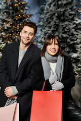 A couple embraces the holiday spirit near snow-laden pine trees, dressed warmly and sharing smiles.