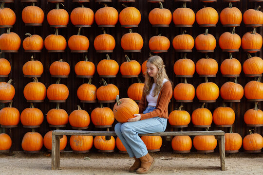 blonde teenage girl sitting on a bench holding a pumpkin in front of vibrant wall of pumpkins