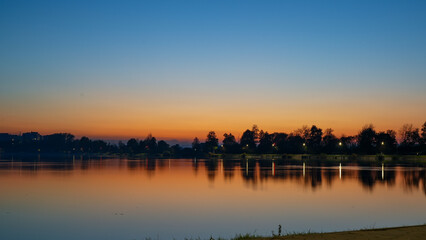 Naklejka premium Romantic sunset on a lake. The sun is barely visible because of the silhouettes of the trees. Dawn on the lake, the sun comes out of the trees. autumn landscape in the evening.