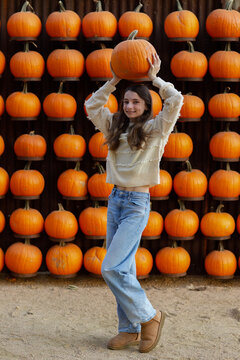 girl carrying a pumpkin above her head in front of a wall of pumpkins