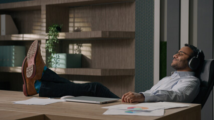 Calm relaxed American businessman Indian man Arabian worker entrepreneur in headphones listen music song in sound app legs on table office enjoy audio relaxing resting work break finish tranquil relax