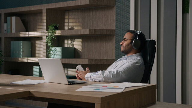 Calm relaxed American businessman Indian man Arabian worker entrepreneur in headphones listen music song in sound app legs on table office enjoy audio relaxing resting work break finish tranquil relax