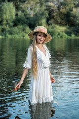 Happy little girl with long blonde hair in a white dress and straw hat in the river