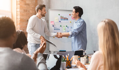 Obraz premium Young guys colleagues shaking each other hands during presentation, international colleagues apllausing, office interior