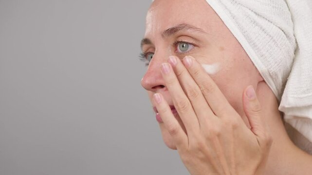 Skincare advertising concept. Calm beautiful woman wearing towel on head on studio gray background, applying facial moisturizer. Morning beauty routine. Preparation for make up. Copy space.