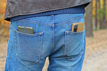 two different in plastic case modern orange with glass camera and display electronic portable mobile phones in man's back pockets blue stylish fashion jeans and leather brown jacket daytime in forest