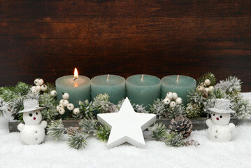 Adventsdekoration zum ersten Advent. Grüne Kerzen vor einer winterlichen Kulisse mit Schnee, Tannenzweigen, zwei Schneemännern und Platz für Text.