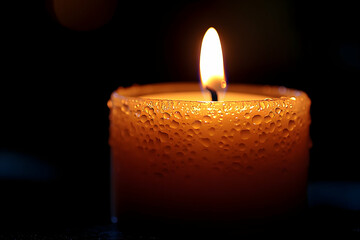 A close-up of a glowing candle, featuring a warm flame and droplets of wax on its surface, creating a cozy ambiance against a dark background.