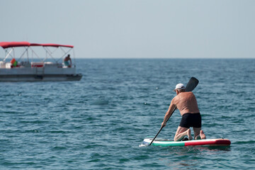 An elderly man operating a sap-board with a paddle on the sea. A boat is in the background.