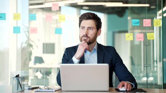 Businessman thoughtfully working in modern office with laptop. Male professional wearing formal suit focused on planning and strategy development. Concept of business decision making and innovation