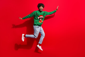 Full length photo of handsome young guy jump have fun walk dressed green christmas sweater celebrate winter holidays on red background