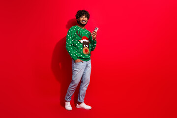 Full size photo of pretty young male hold device wear green christmas sweater masquerade theme party on red color background