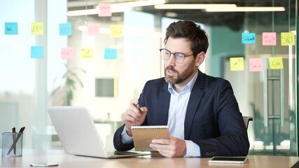 Businessman watching video call online conference taking notes looking at laptop computer screen sitting at workplace in office. Male listening remote business training or course talking with a tutor