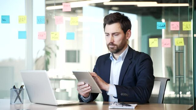 Portrait of smiling businessman using digital tablet sitting at desk at workplace in business office. Confident and professional male employee working indoors. Headshot manager poses looking at camera