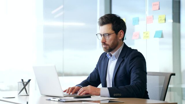 Businessman in a formal suit works on laptop computer sitting at workplace in business office. Focused serious manager in glasses banking, typing texting a client, chatting online. busy with a project