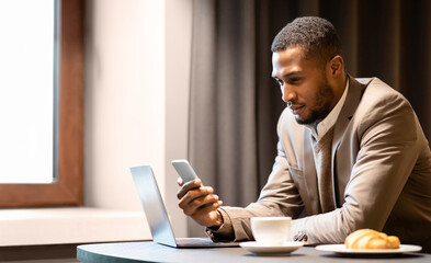 Working Anywhere. Concentrated young black man using his cellphone and laptop spending time in restaurant, copyspace