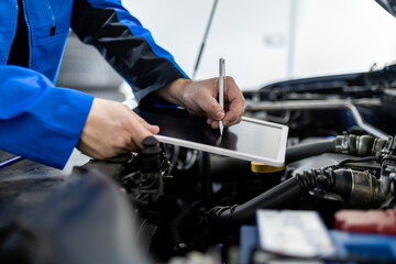 A mechanic inspects a vehicle while taking notes on a tablet in a repair shop during the day
