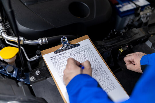 Mechanic reviewing maintenance logs while inspecting a vehicle’s engine in an auto workshop during daylight hours