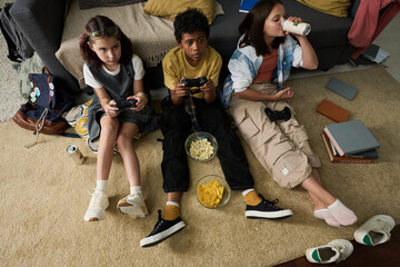 Kids sitting on carpet with game controllers, snacks, and drinks while enjoying video games in cozy...