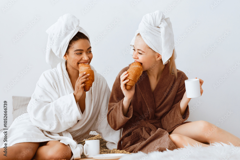 Wall mural happy multiethnic girlfriends in bathrobes and towels on heads sitting on bed and having breakfast a