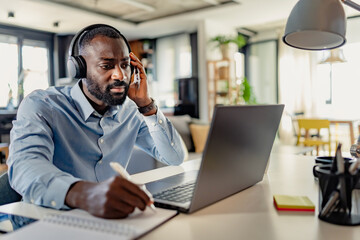 African-American man wearing headphones, taking notes while working on a laptop from home, focused and engaged in a modern workspace.