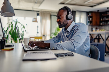 Obraz premium Focused man wearing headphones, working on his laptop in a modern, well-lit home office environment.