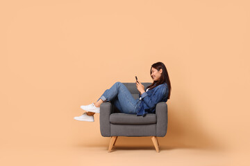 Smiling woman with smartphone sitting in armchair on beige background